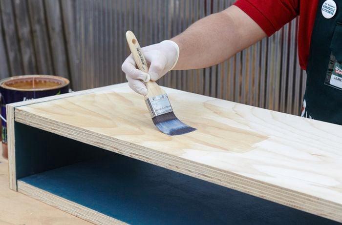A person painting clear sealer onto a plywood box using a paint brush