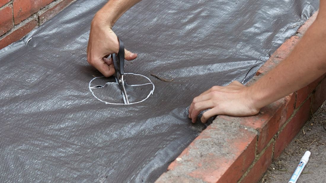 A hole being cut through installed weed matting with garden shears