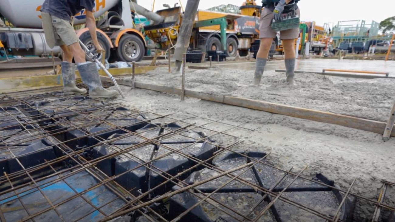Workers in gumboots pour and screed wet concrete over Biax pods and steel reinforcement. A pump hose delivers the concrete, with construction trucks in the background