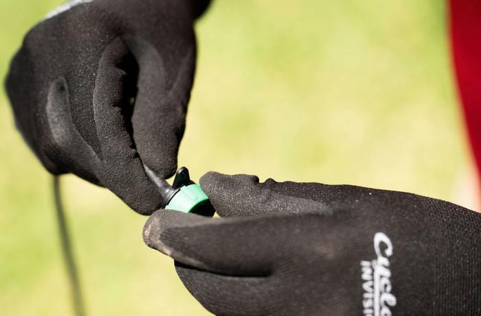 A person wearing black gloves is holding a dripper irrigation component, attaching a green and black emitter to a tube.