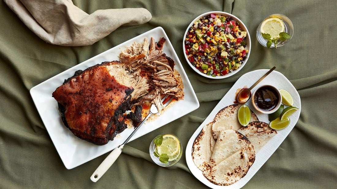 Smoked pulled pork on a platter with a serving fork, a bowl of cowboy caviar and a plate of tortillas with lime wedges and barbecue sauce