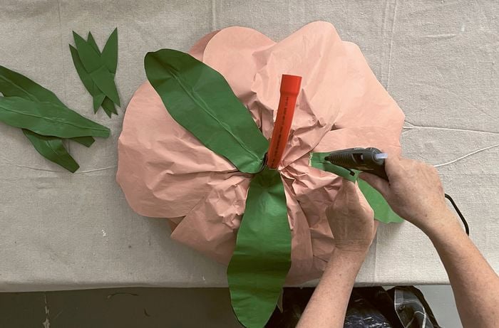 Hands using a hot-glue gun to attach green-painted paper leaves to a large, peach-coloured paper flower with an orange conduit stem.