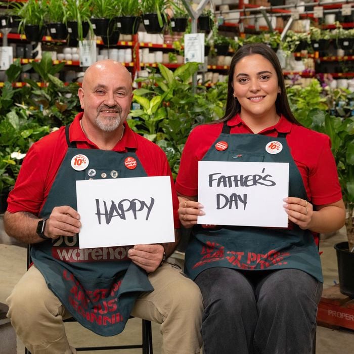 Two Bunnings team members seated in the indoor garden section of a store, surrounded by shelves of potted plants and hanging greenery. Each person holds a white sign, together displaying the message “HAPPY FATHER’S DAY.” Both are wearing red shirts and dark green Bunnings aprons.