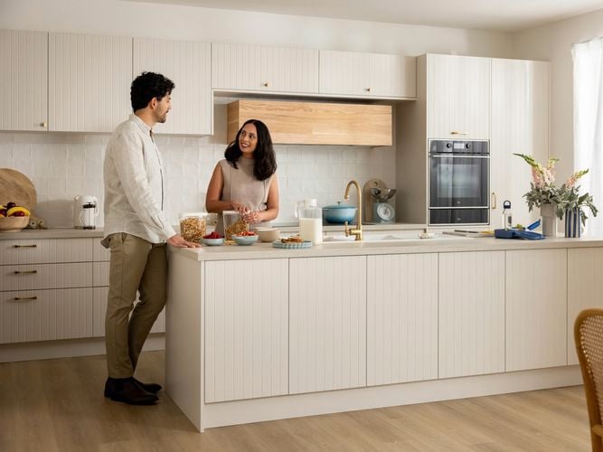 Couple in early 40's standing in a vanilla coloured kitchen island smiling at each other as they prepare breakfast.