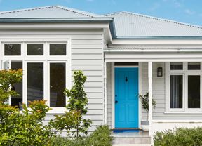 A white house with a bright blue front door, surrounded by green shrubs and a grey-tiled porch