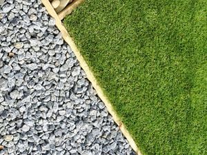 Top-down view of a landscaped area with beige stones, gray gravel, and green grass with a palm tree, separated by wooden or stone borders."