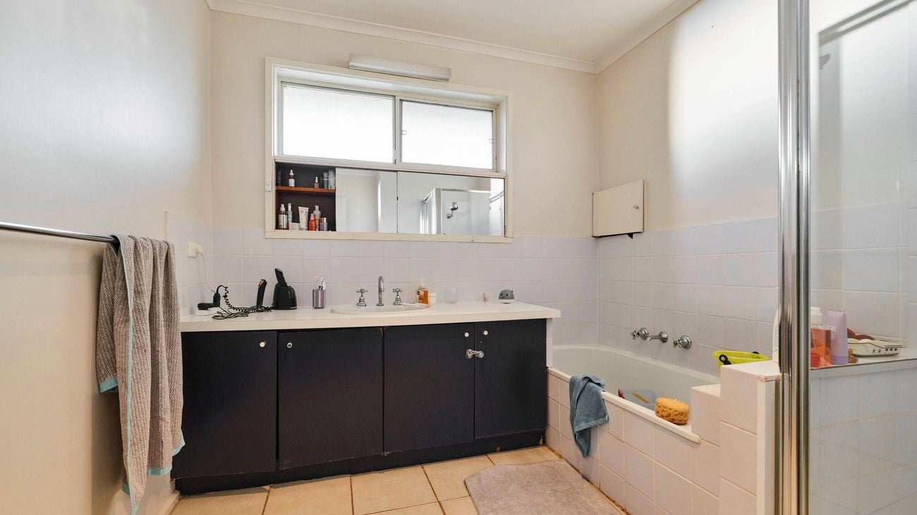   Bathroom with a dark vanity cabinet and white benchtop, featuring a central sink beneath a large mirror and window, a tiled bathtub with chrome taps and a sponge on the edge to the right, a glass shower screen, beige tiled flooring with a bath mat, and towels and toiletries arranged around the space.