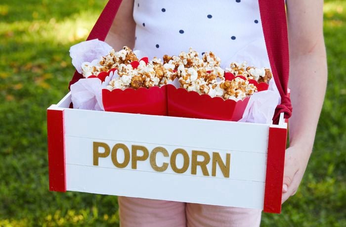  Child holding a painted wooden crate tray labelled ‘popcorn’ with buckets of popcorn inside.