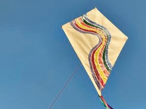 Beige diamond-shaped kite with retro rainbow stripes, attached to a red line, flying against a clear blue sky.