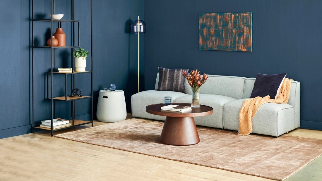 Lounge room with brown shag rug, white cement side table, blue feature wall, decorative shelf, white lounge and brown coffee table.