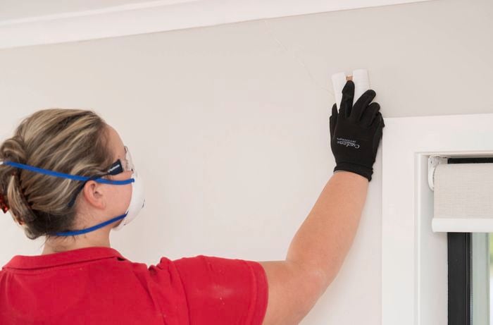 A person wearing a mask and gloves is sanding a patched crack on a wall to remove any bumps or ridges near a window. 