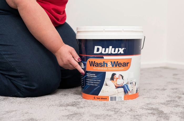 A person in a red T shirt is kneeling on a carpet and pointing at a Dulux Wash & Wear paint bucket