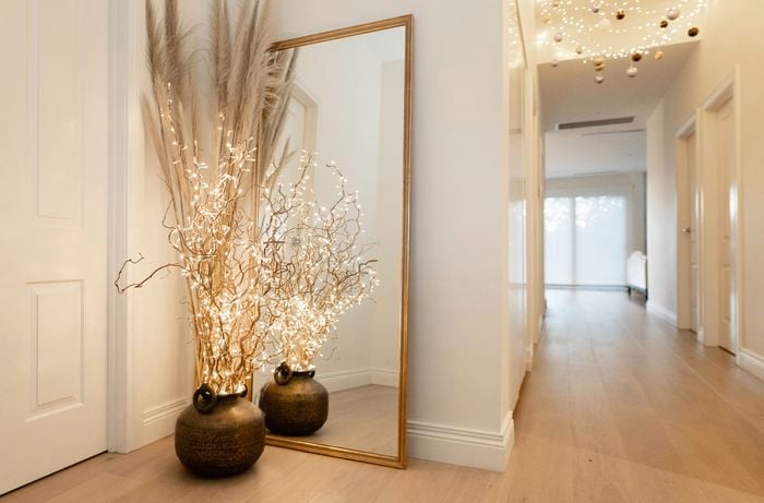 A vase with branches and lights is placed in front of a large mirror in a hallway, with string lights hanging from the ceiling
