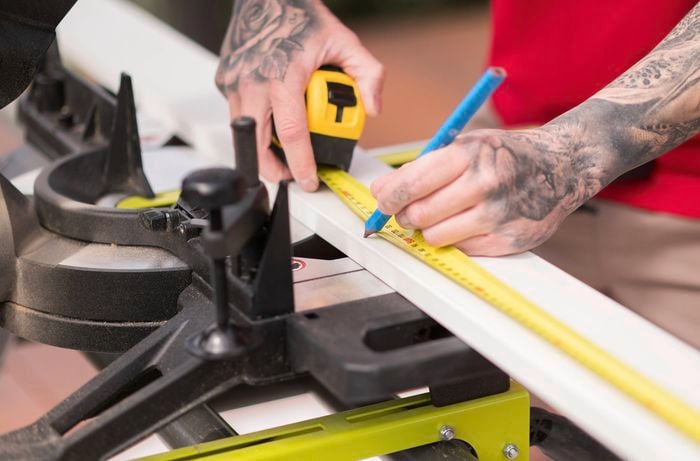 A person with tattooed arms is marking the measurements on a skirting board to indicate where it needs to be cut. 