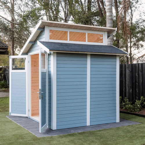 Modern light blue garden shed with asymmetrical roof and windows, viewed from the rear corner in a landscaped backyard.
