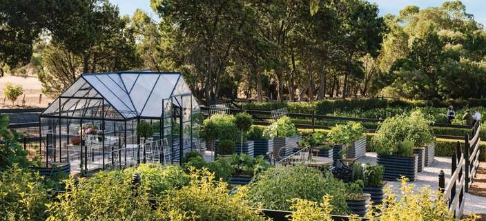 A greenhouse in a lush garden with raised vegetable beds and trees.