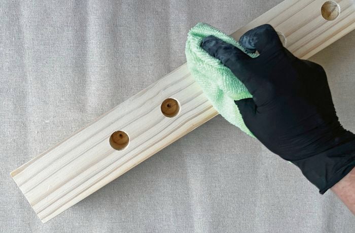 A hand wearing a black glove wipes a white painted timber block with a green cloth, on a drop sheet