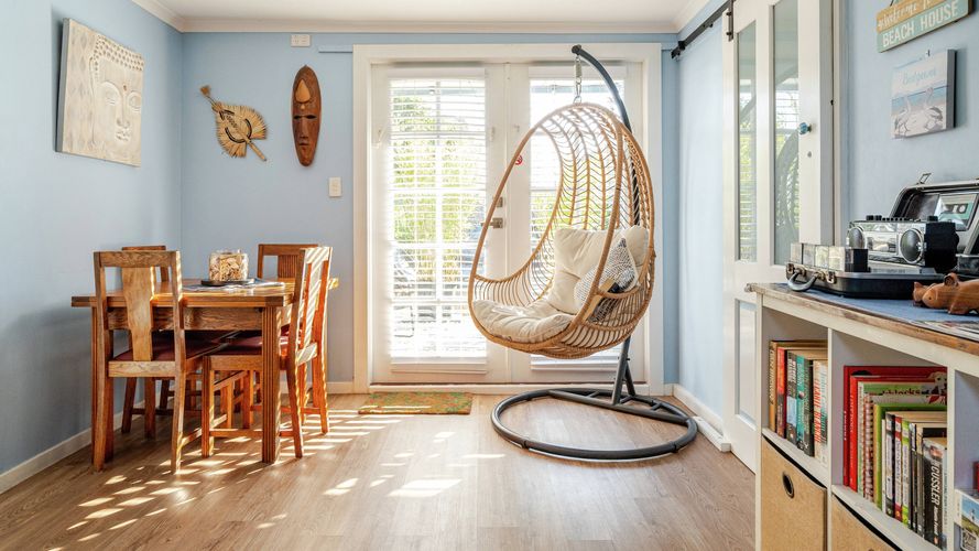 Light-filled dining and living area with pale blue walls, timber dining table, hanging wicker chair, bookshelves and French doors opening to the outdoors.