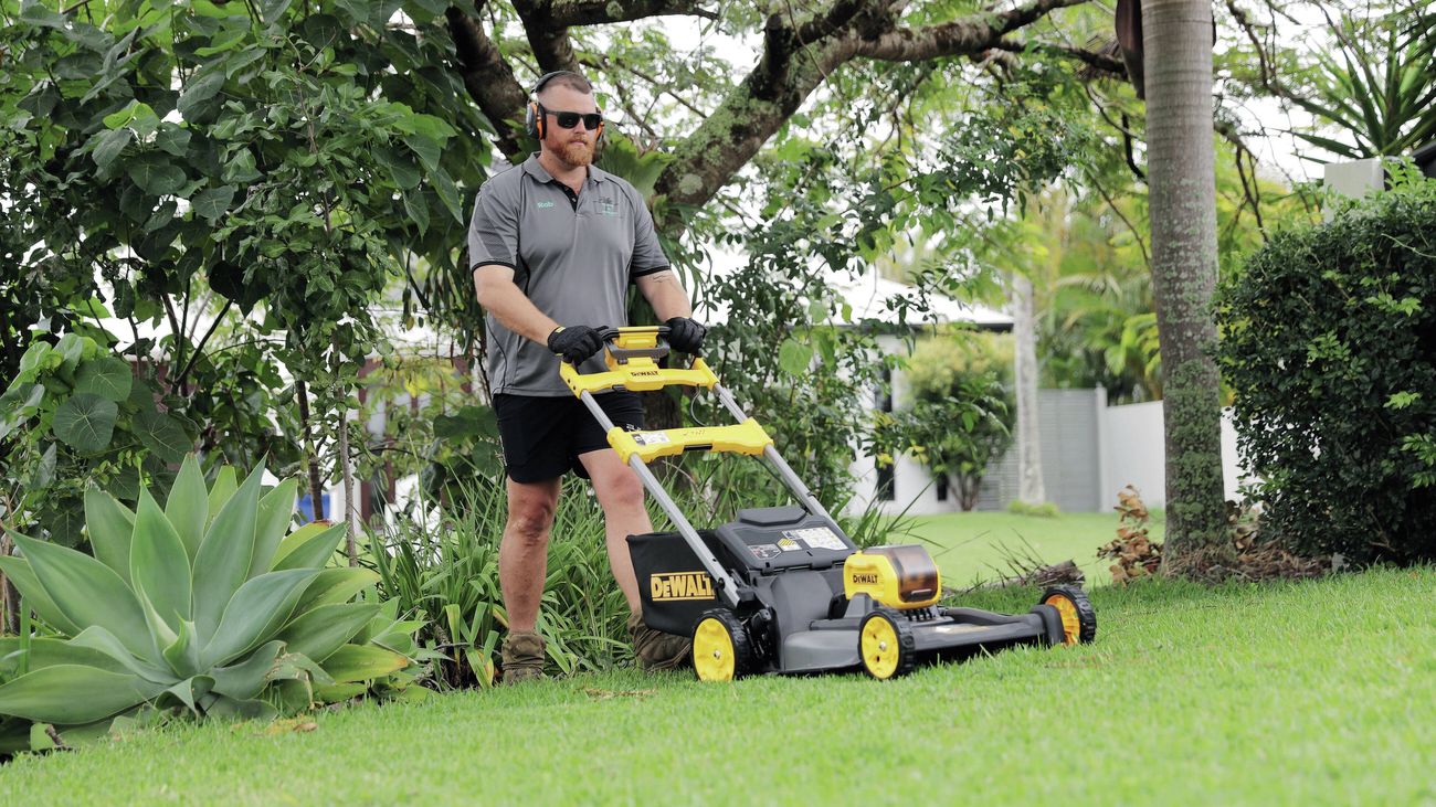 Person pushing a lawn mower across a green lawn in a landscaped front yard.