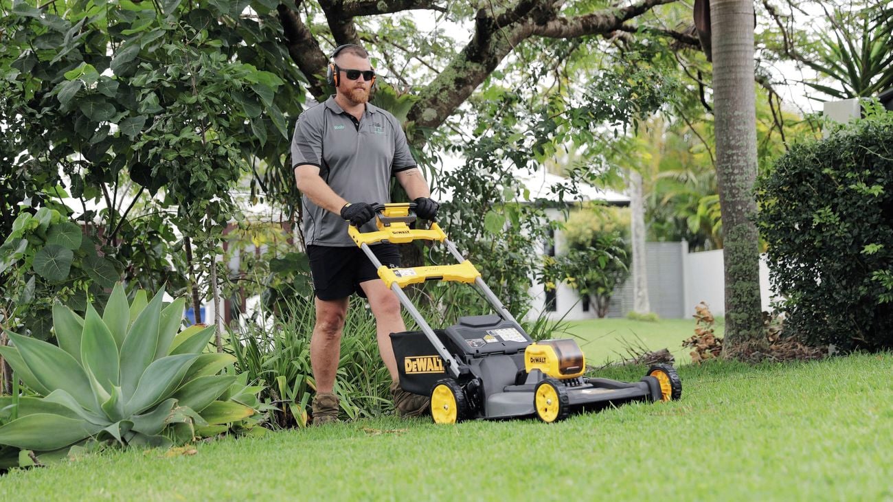 Person pushing a lawn mower across a green lawn in a landscaped front yard.