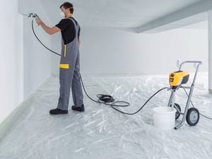 Person in protective gear using a paint sprayer to paint a room, with plastic sheeting on the floor and equipment nearby.