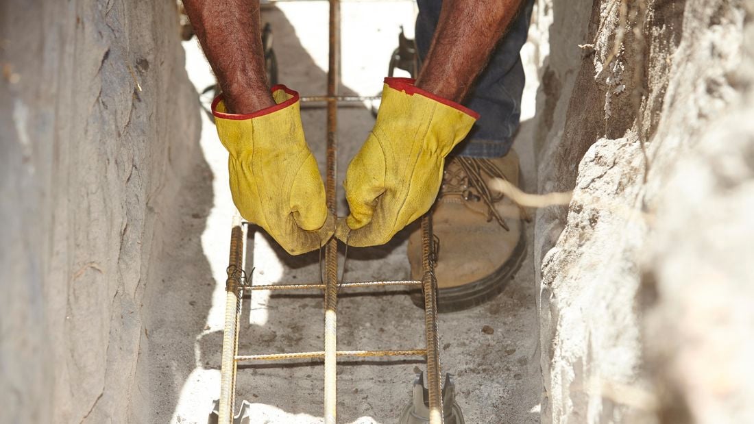 Person tying wire around pieces of reinforced steel in a dirt trench.