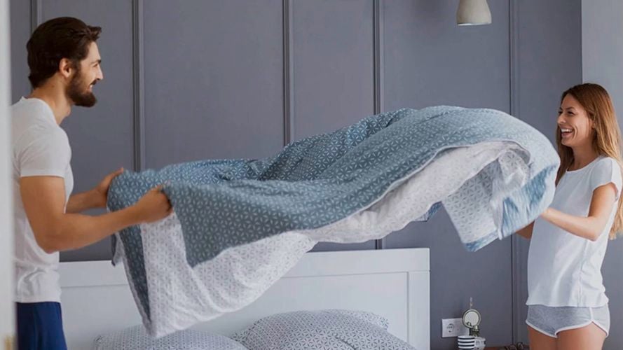 Two people adjusting a patterned blanket on a bed in a modern room.