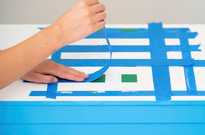 A person is re-taping the painted squares on the toy box lid to refine the grid pattern for a DIY games board design.
