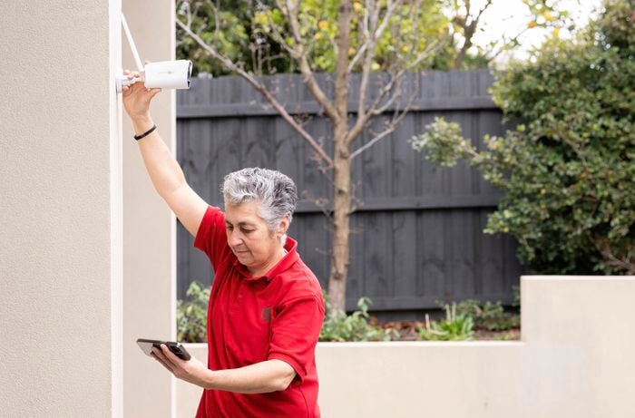 A Bunnings Team Member is installing a Swann security camera on a wall while using a smartphone to check the best signal placement.