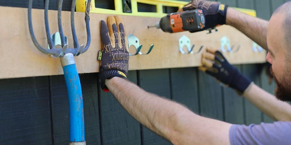 Person attaching a garden tool holder to a timber paling wall with a drill and wearing gloves