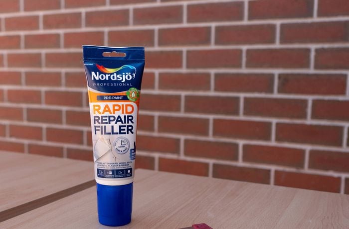 A tube of Rapid repair wood filler and a putty knife are placed on a wooden surface to fill the existing holes.