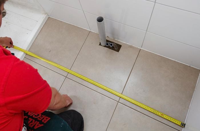 A person using a measuring tape to mark out the position of a vanity unit on a tiled floor 