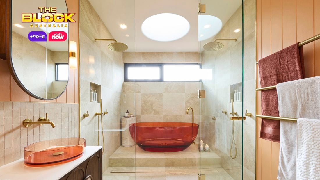 Modern bathroom with amber tub in glass shower, beige tiles, dual overhead showerheads, amber basin with round mirror, and gold towel rack. 