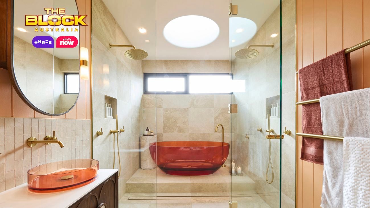 Modern bathroom with amber tub in glass shower, beige tiles, dual overhead showerheads, amber basin with round mirror, and gold towel rack. 