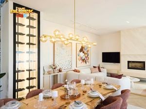 A modern living and dining area featuring a dining table set with plates, glasses, and candles, surrounded by six chairs. A contemporary chandelier with spherical lights hangs above. In the background, a white sofa with colorful cushions sits beneath two large abstract artworks. A fireplace with a TV above it is on the right, and a tall wine rack filled with bottles is on the left. 