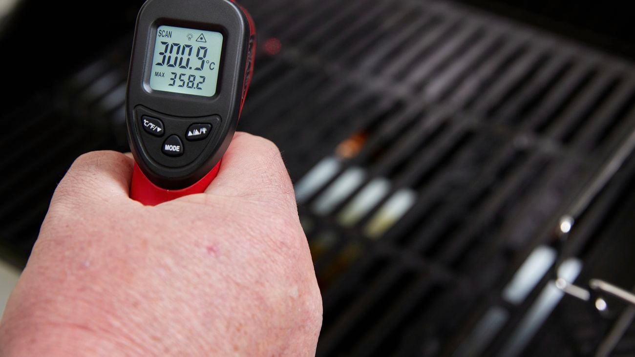Hand holds a red and black infrared thermometer at a BBQ with the temperature showing on its screen
