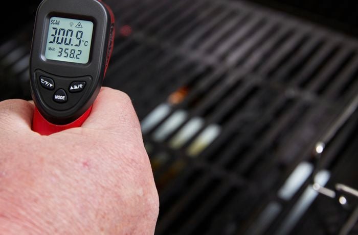 Hand holds a red and black infrared thermometer at a BBQ with the temperature showing on its screen