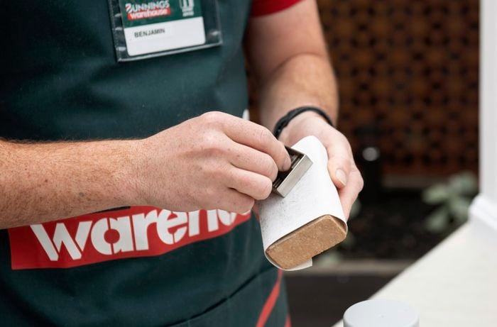The surface of a wooden drawer handle being sanded down with sandpaper wrapped around a scrap piece of wood