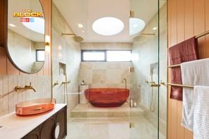 Modern bathroom with amber tub in glass shower, beige tiles, dual overhead showerheads, amber basin with round mirror, and gold towel rack. 