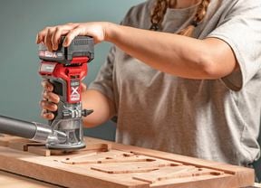 A woman uses a red Ozito PXC router on a wooden board with cut-out shapes.