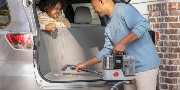 A man and child seated in the rear of a car, engrossed in conversation, cleaning the boot upholstery with a Hoover Clean Slate Pot Washer.