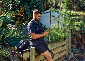 A landscaper sits on a timber retaining wall outside looking at his phone