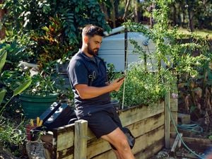 A landscaper sits on a timber retaining wall outside looking at his phone