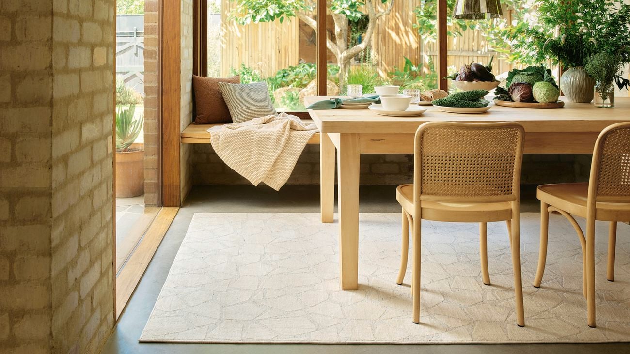 Dining area with a wooden table and cane-backed chairs on a patterned rug. A built-in wooden bench with cushions and a blanket sits by a large window overlooking a garden.]