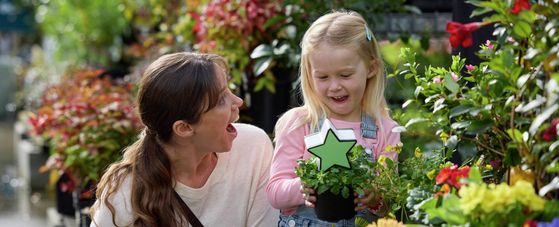 Customer browsing flowering plants in a garden centre with a child holding a potted flowering plant.