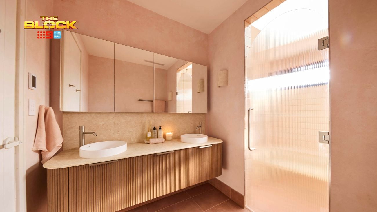 Modern bathroom with double round sinks, chrome faucets, large mirror, frosted glass shower door, brown tiled floor, and light pink walls. 