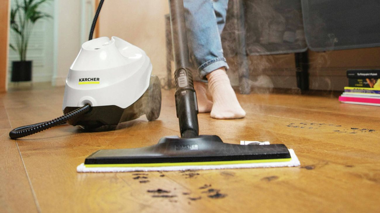 Person steam-cleaning a wooden floor wearing jeans and socks with a white Kärcher steam cleaner.