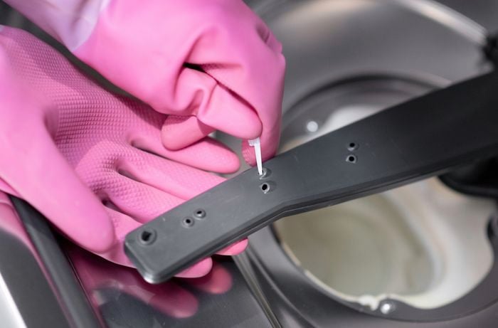 A hand wearing pink gloves is using a toothpick to clean the spray arms of the dishwasher.