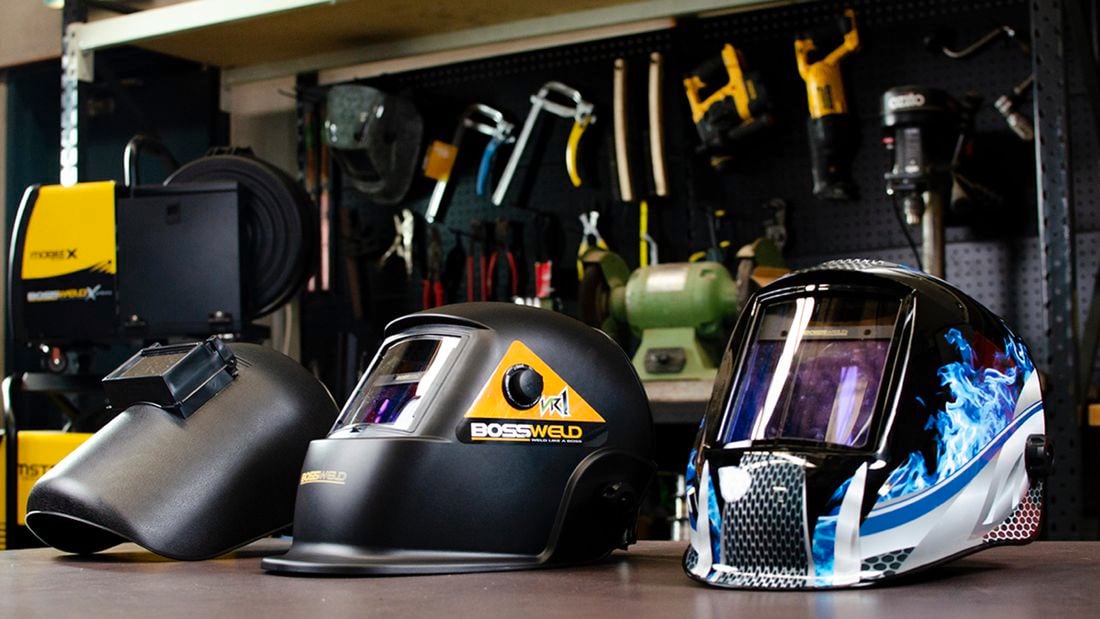 Three different sorts of welding helmets on  a bench in a workshop.