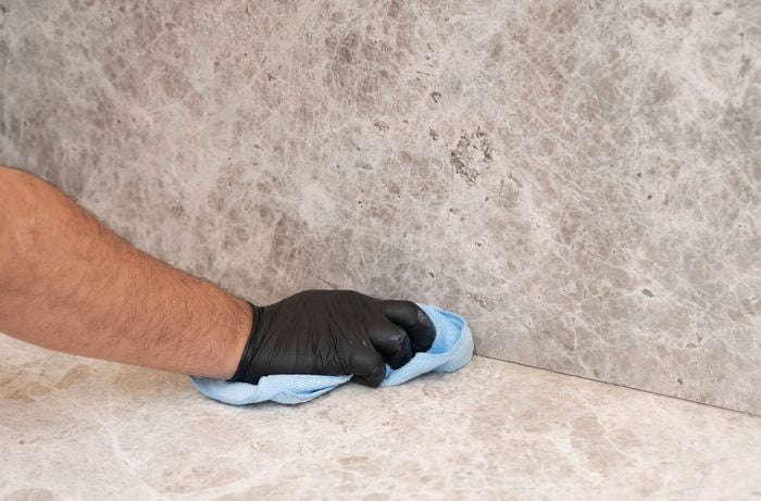 Hand wearing a black glove wipes the junction between a stone-look benchtop and matching splashback with a blue microfiber cloth to clean the surface before sealing.